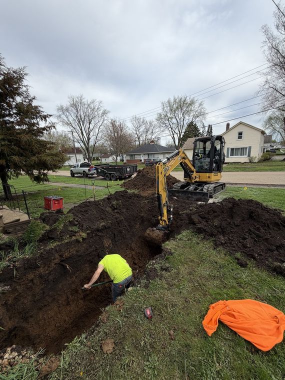Mini excavator digging an open trench across a residential side yard with a worker in the trench