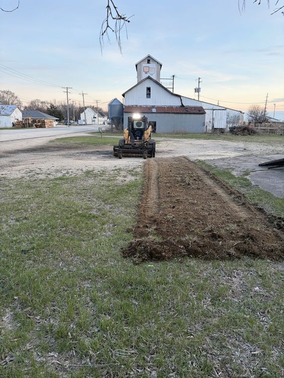 Skid steer grading a long strip of bare soil beside a rural outbuilding